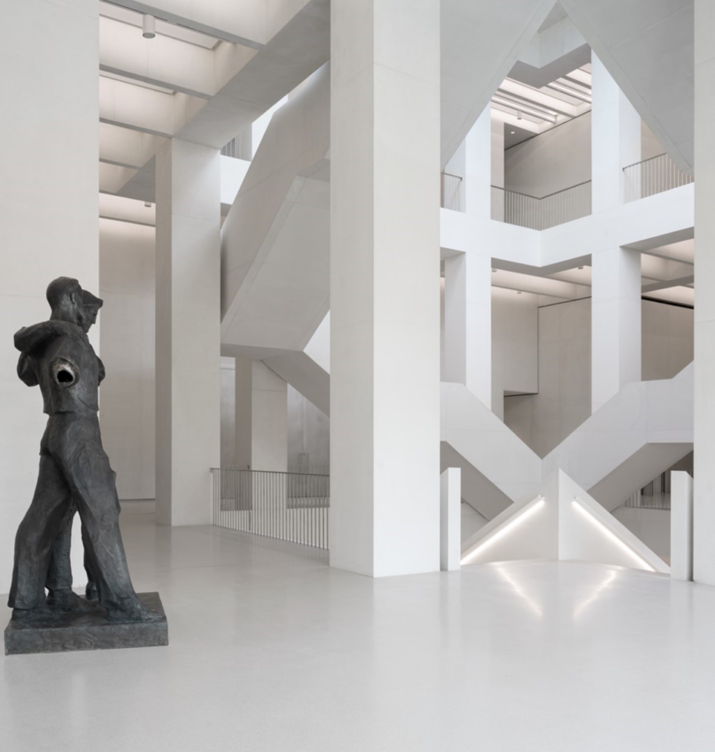 Witte atriumhal van MSN Warschau met zigzaggende trappen en een bronzen sculptuur van twee omstrengelde figuren.