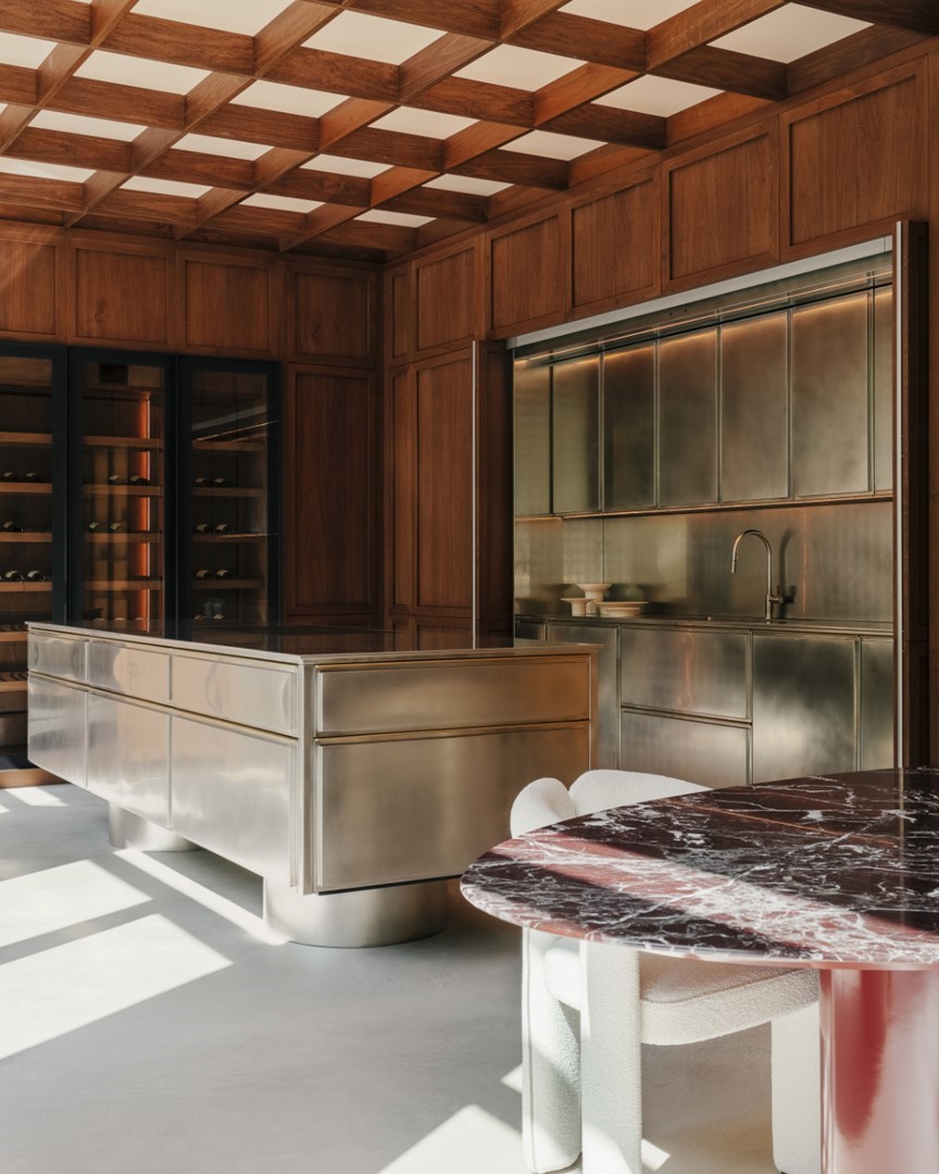 Moderne luxe keuken met groot stalen kookeiland, ingebouwde wijnkast, messingkleurige kastenwand en ronde eettafel met rood marmeren blad