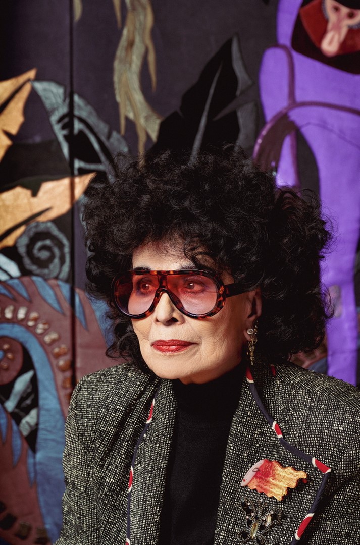 Portret van modeontwerper Fong-Leng (1937) met grote zonnebril, rood opgemaakte lippen en zwarte krullen, poserend voor een kleurrijk wandkleed met abstracte patronen.