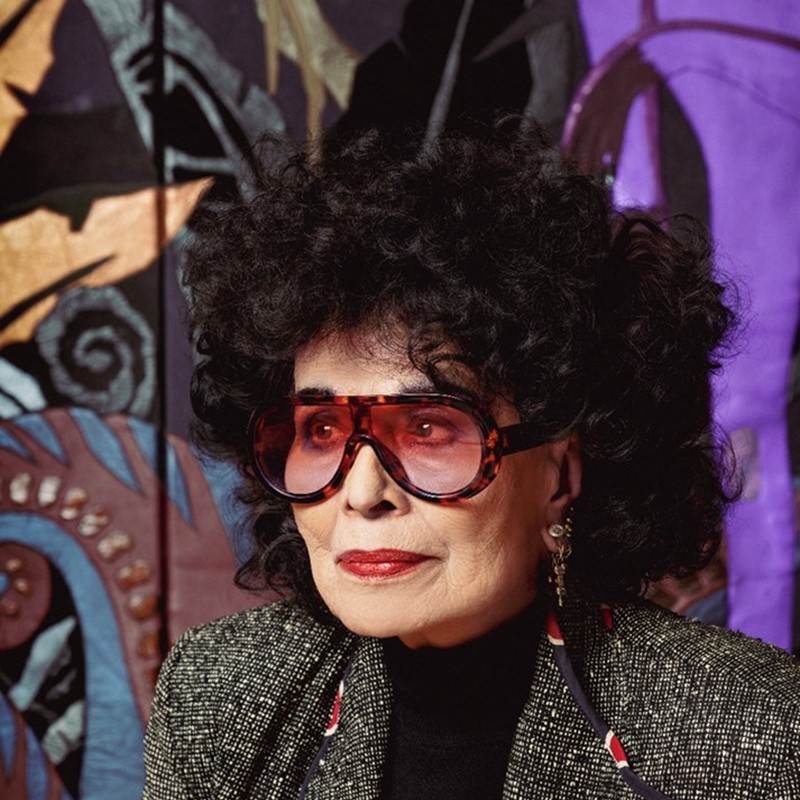 Portret van modeontwerper Fong-Leng (1937) met grote zonnebril, rood opgemaakte lippen en zwarte krullen, poserend voor een kleurrijk wandkleed met abstracte patronen.