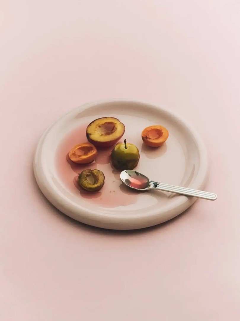 Zachtroze bord met pruimen, abrikozen en lepel op een lichte, minimalistische achtergrond