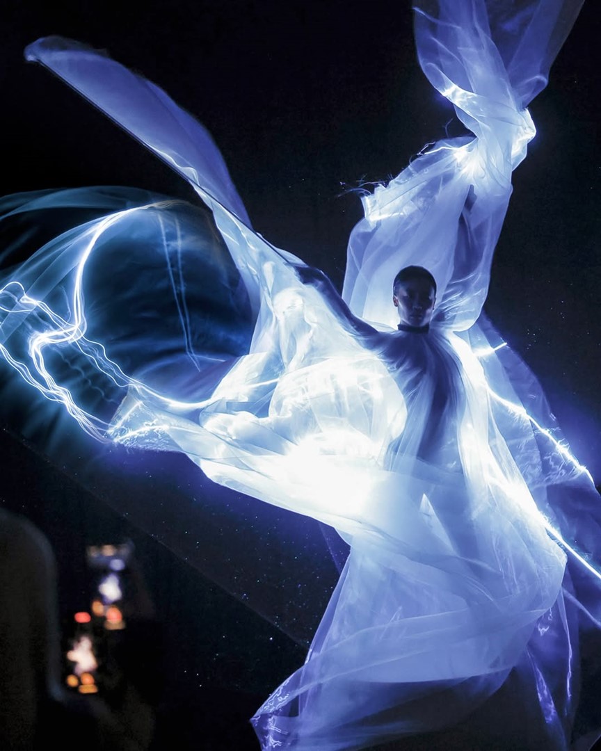 Mode-installatie uit de tentoonstelling Iris van Herpen: Sculpting the Senses in Kunsthal Rotterdam, met een model omhuld door transparante, lichtgevende stoffen die golvend door de ruimte bewegen en een futuristisch, etherisch tafereel oproepen.