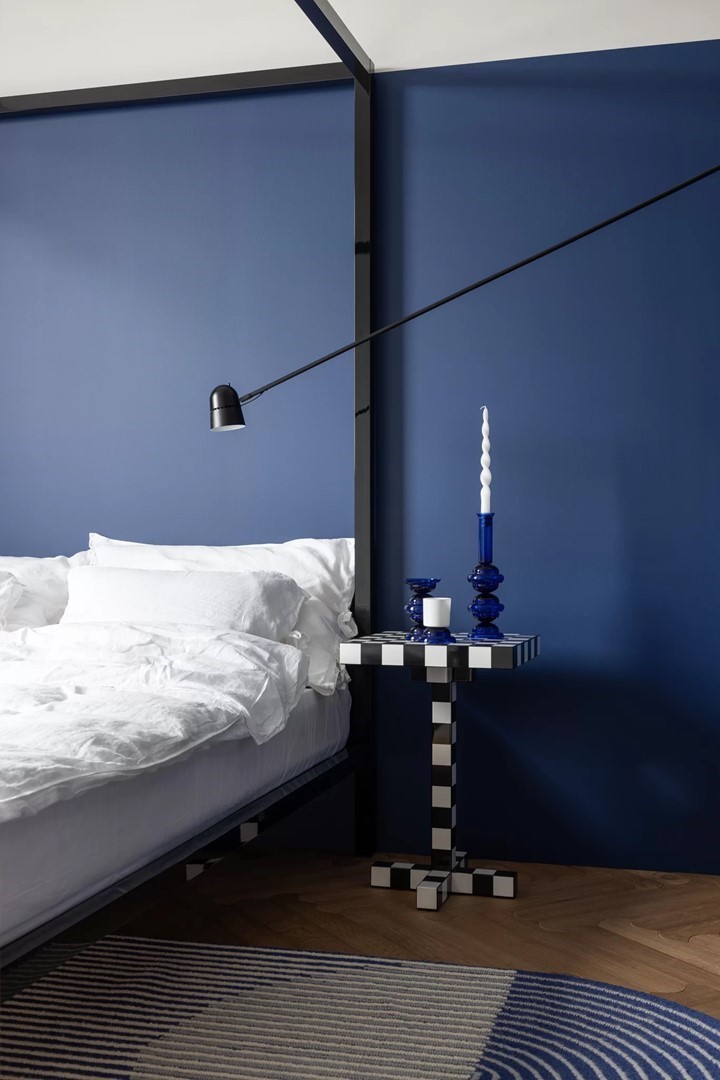 Slaapkamer met blauwe muur, wit beddengoed en de Chess Table van Moooi als nachtkastje, gestyled met blauwe kandelaar.