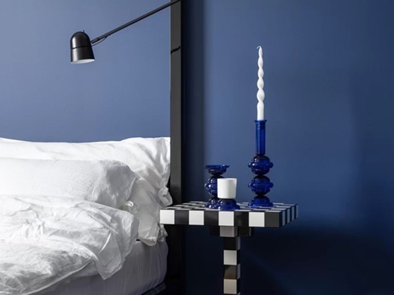 Slaapkamer met blauwe muur, wit beddengoed en de Chess Table van Moooi als nachtkastje, gestyled met blauwe kandelaar.