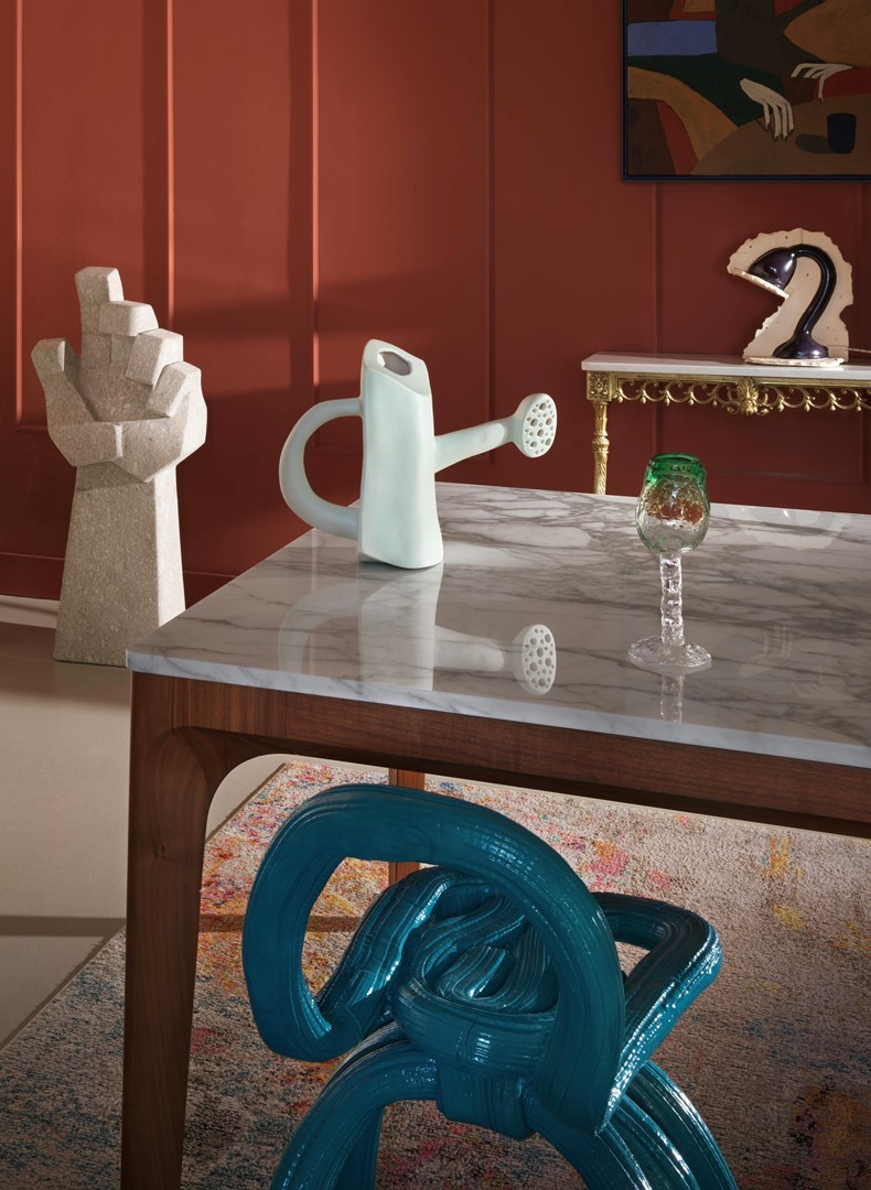 Marmeren eettafel met walnoothouten frame en sculpturale turquoise stoel; op het blad een mintgroene keramische gieter en glas met groene rand. Tegen een roodpanelen muur staan een geometrisch stenen handsculptuur, een verguld console met keramiek en een abstract schilderij—een speelse mix van kunst en design.