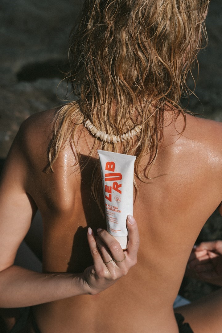 Le Rub All-Day Sunscreen SPF 30 vastgehouden achter de rug bij een rotsachtig strand