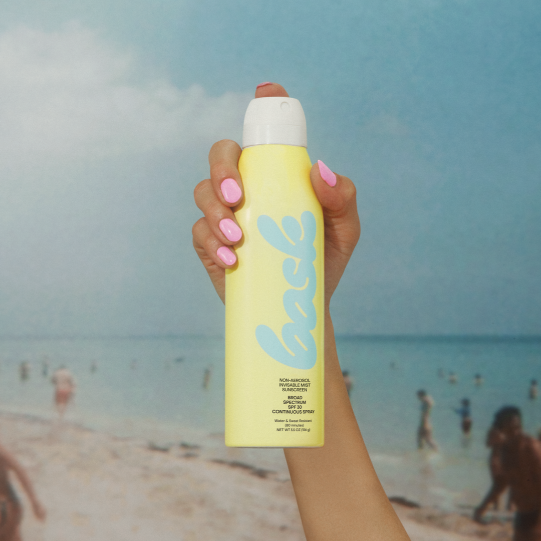 Bask Non-Aerosol Spray SPF 50 in gele fles omhooggehouden tegen een strandachtergrond