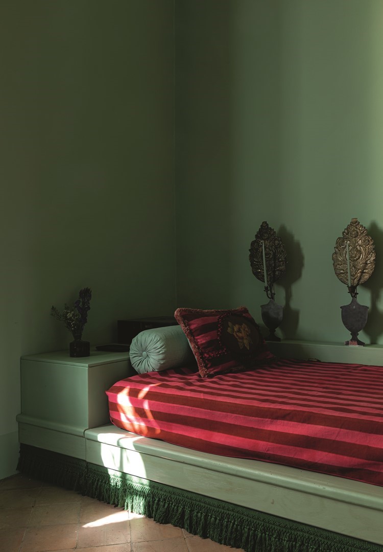 Slaapkamer met saliegroene muren en een ingebouwd bed met rood-roze gestreept matras, ronde mintgroene rolkussen en decoratief sierkussen; naast het bed staan twee klassieke wandornamenten, terwijl zonlicht strepen werpt op de terracottavloer.