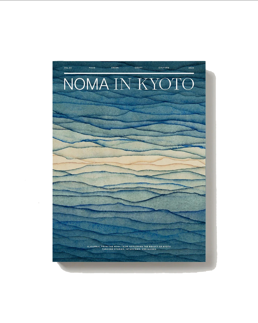 magazinecover met de titel “NOMA IN KYOTO” in grote witte letters bovenaan. De achtergrond bestaat uit gelaagde, golvende aquarelstructuren in verschillende tinten blauw en zacht beige, die doen denken aan water of berglandschappen. Bovenaan staan kleine woorden als “Vol 01”, “Food”, “Drink”, “Craft”, “Culture” en “2023”. Onderaan staat in kleine letters een beschrijving van het tijdschrift als een journal van het Noma-team over Kyoto.