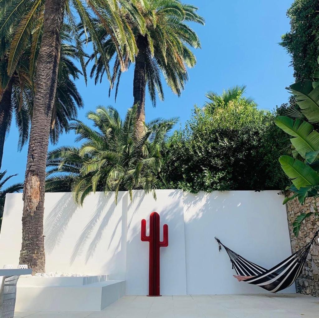 Zonnige patio met witte muren, omringd door hoge palmbomen en weelderig groen. Tegen de muur staat een opvallend rood cactusvormig kunstwerk, terwijl rechts een zwart-witte hangmat is opgehangen.