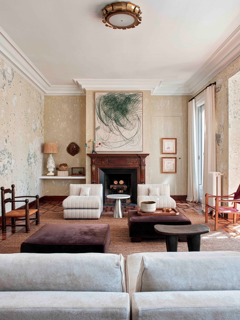 Woonkamer door Quintana Partners met verweerde kalkwanden, houten schouw en abstract groen kunstwerk; lage crèmekleurige fauteuils, bordeaux fluwelen poefs, juten kleed en parket—een mix van vintage en hedendaagse stukken in zacht licht.