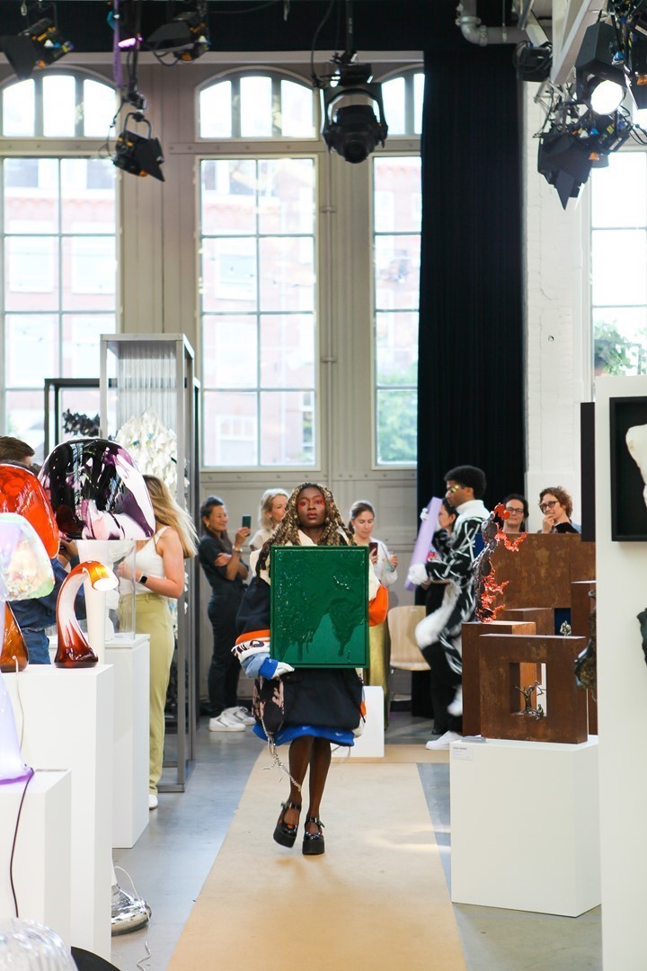 Een kleurrijk, abstract schilderij hangt aan een witte galeriewand. Op de voorgrond staat een onscherpe silhouet van een persoon die naar het kunstwerk kijkt. Het schilderij bevat zachte blauwtinten, geel en oranje accenten en organische vormen.