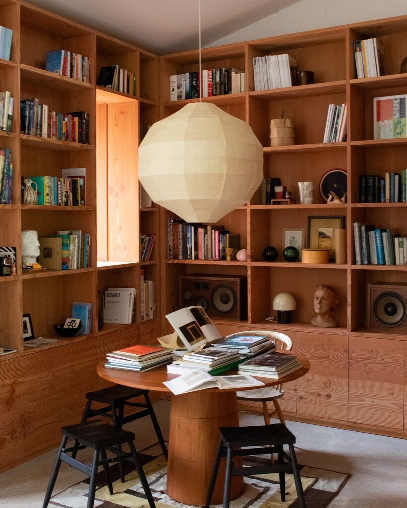 Houten boekenkastwand met open planken gevuld met boeken en objecten, een ronde houten tafel met stapels kunstboeken en zwarte krukjes eromheen, verlicht door een grote, ronde papieren hanglamp.