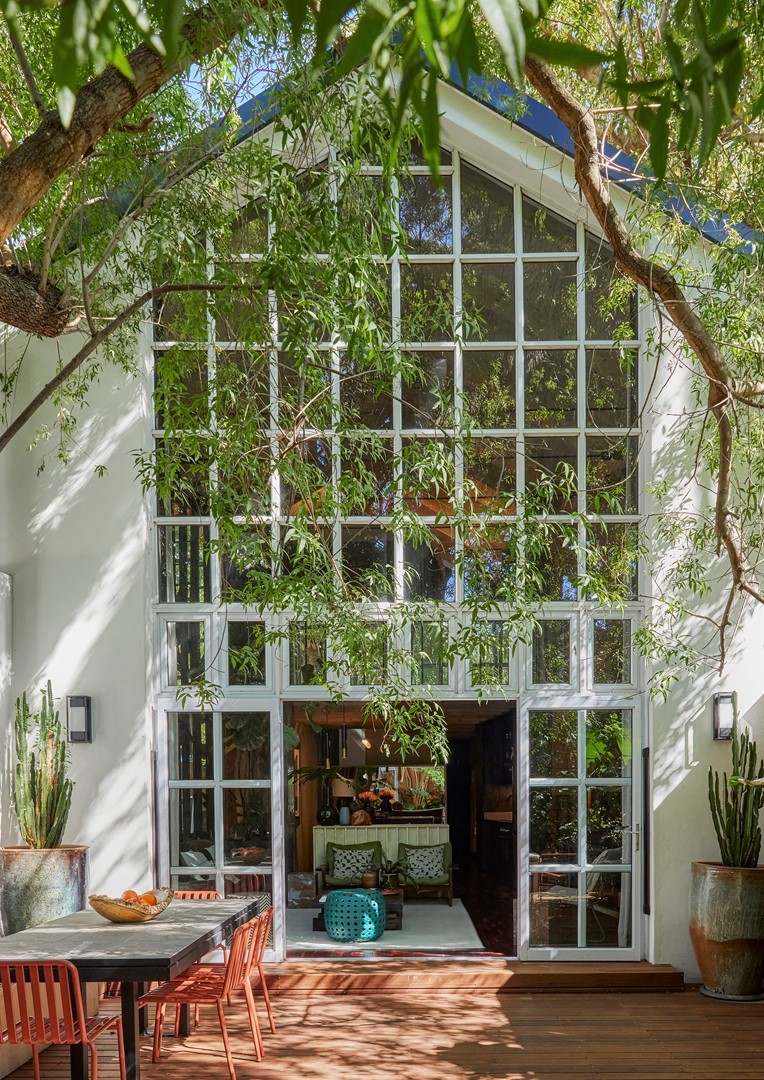 Moderne woning met een hoge glazen pui, omgeven door bomen en groen. De openslaande deuren verbinden een houten terras met een eettafel en stoelen met een sfeervol interieur, zichtbaar door het raam.