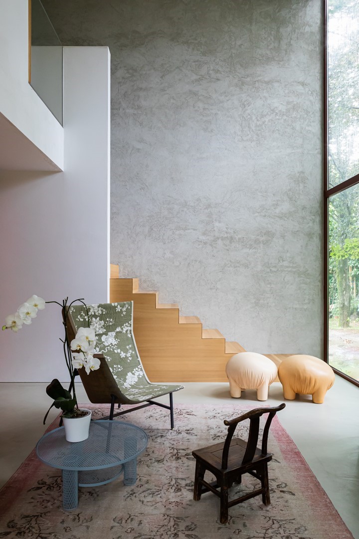 Dubbelhoge living met grote raampartij op bos, grijze stuucwand en eiken trap in ‘zigzag’-profiel; groene loungefauteuil met bloemenprint, blauw metalen salontafel met orchidee, klein antiek stoeltje en twee sculpturale, zandkleurige poefs.