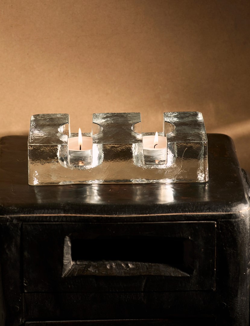 Scandinavische glazen kandelaar uit de jaren 60, uitgevoerd in massief helder glas met twee brandende waxinelichtjes, geplaatst op een donker houten meubel tegen een warme, neutrale achtergrond.