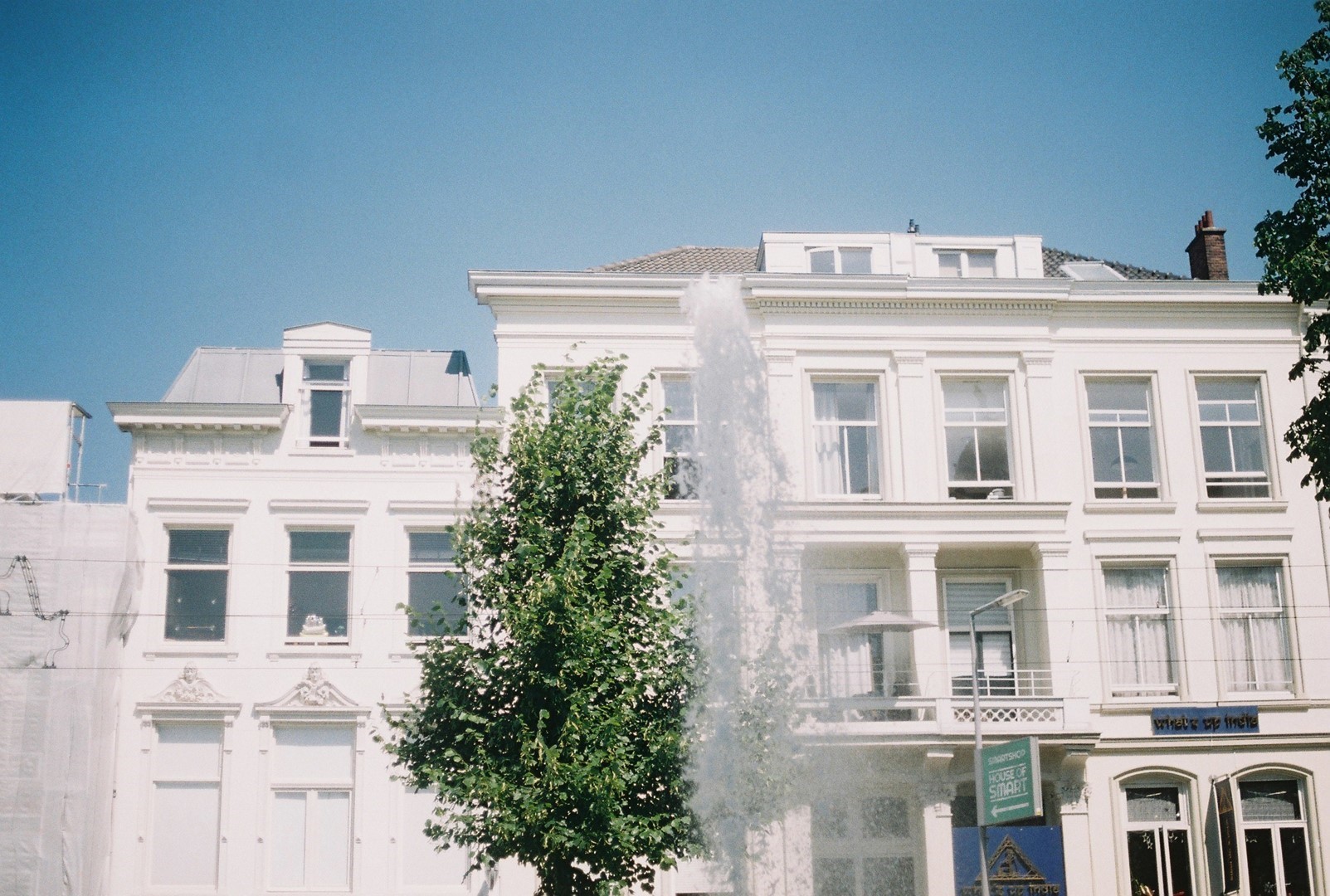 Fontein spuit water omhoog voor een monumentaal wit herenhuis op een zonnige dag, met blauwe lucht en groene bomen op de voorgrond.