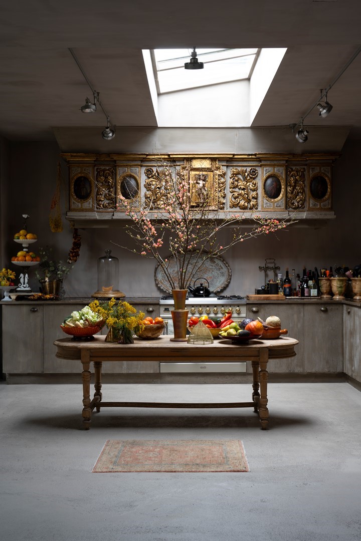 Een sfeervolle keuken met een rustieke houten tafel in het midden, gedekt met schalen vol fruit en een vaas met bloesemtakken. Boven het fornuis hangt een rijk versierde, antieke schouw met gouden details en kleine schilderijen. Het geheel wordt verlicht door een groot dakraam, terwijl keukengerei en flessen langs het aanrecht zijn opgesteld.