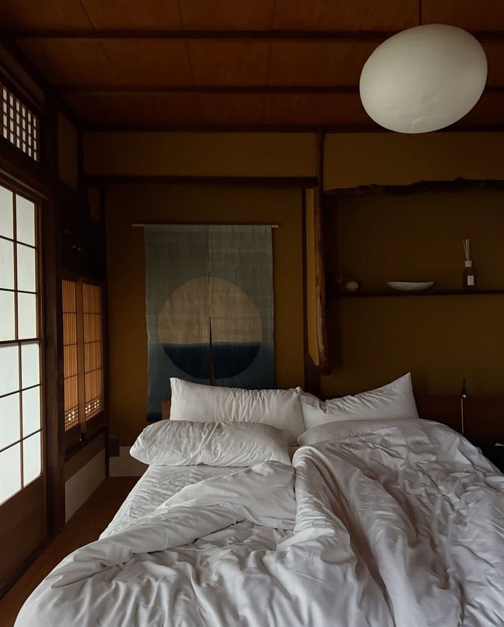 Een sfeervolle Japanse slaapkamer met houten wanden en shoji-schuifdeuren, een laag bed met witte, rommelige dekens en kussens, een ronde papieren plafondlamp en een wanddoek met een abstracte zon boven een donkerblauwe horizon.
