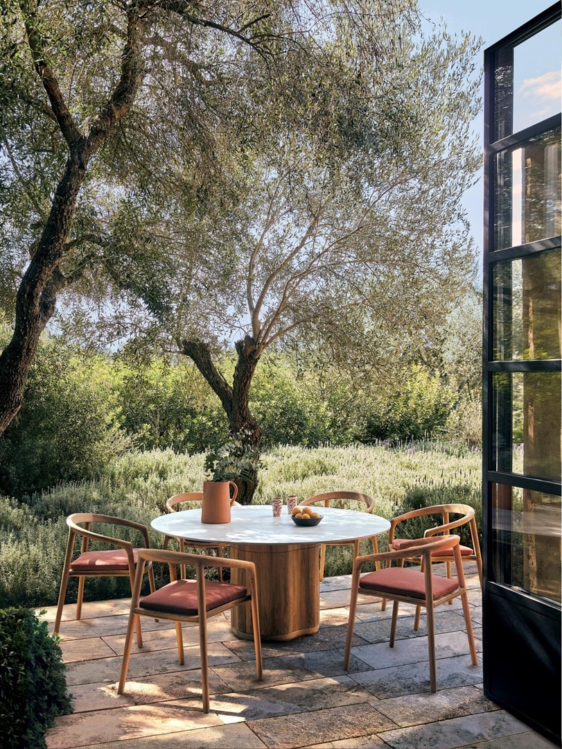 Ronde eettafel met houten stoelen op een zonnig terras, omgeven door olijfbomen en weelderige beplanting. Op de tafel staan een karaf, kopjes en een schaal met fruit, terwijl een open glazen deur zicht geeft op de tuin.