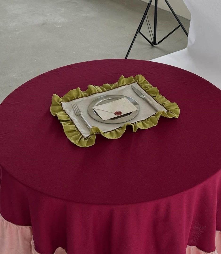 Een ronde tafel met een dieprood tafelkleed. In het midden ligt een beige placemat met een groene gerimpelde rand. Daarop staan een bord met een gevouwen envelop met lakzegel, een vork en een mes.