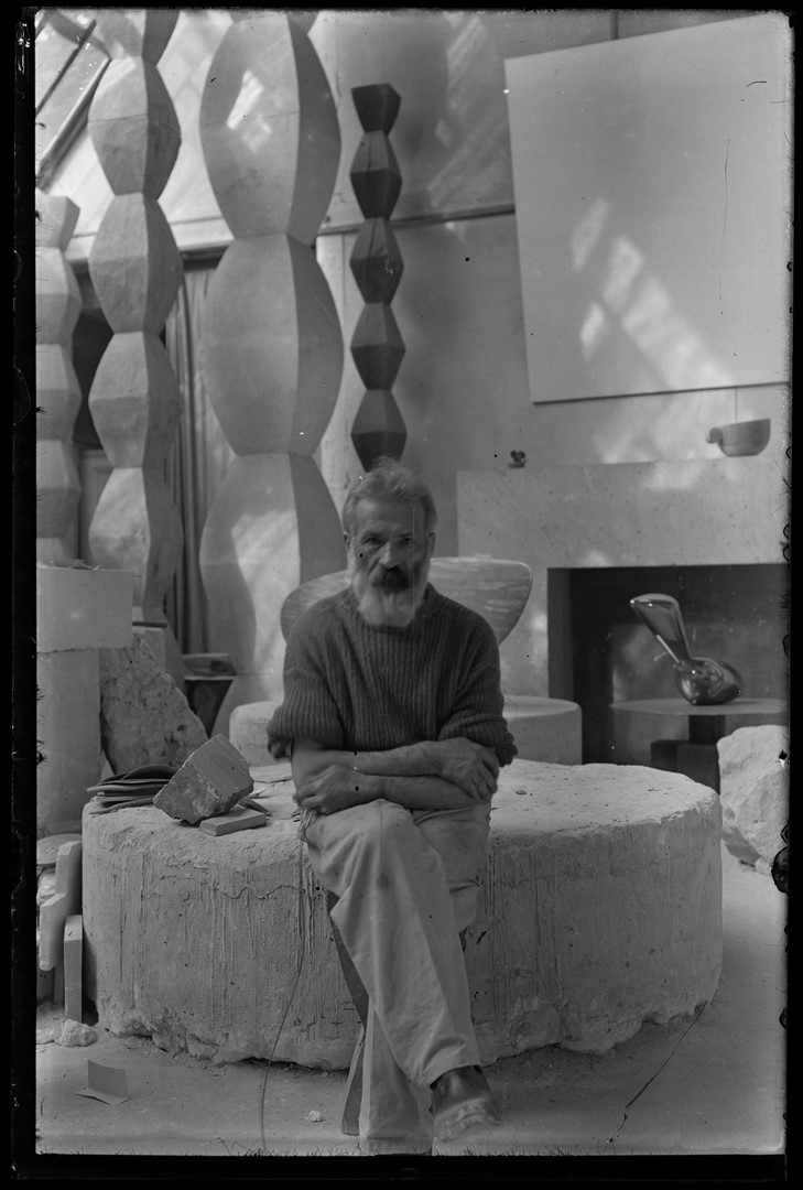 Zwart-witfoto van Constantin Brancusi in zijn atelier in Parijs, zittend voor zijn sculpturen. Te zien in de tentoonstelling Brancusi – The Birth of Modern Sculpture in H’ART Museum Amsterdam, georganiseerd in samenwerking met Centre Pompidou.