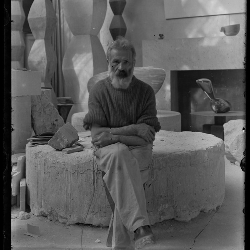 Zwart-witfoto van Constantin Brancusi in zijn atelier in Parijs, zittend voor zijn sculpturen. Te zien in de tentoonstelling Brancusi – The Birth of Modern Sculpture in H’ART Museum Amsterdam, georganiseerd in samenwerking met Centre Pompidou.